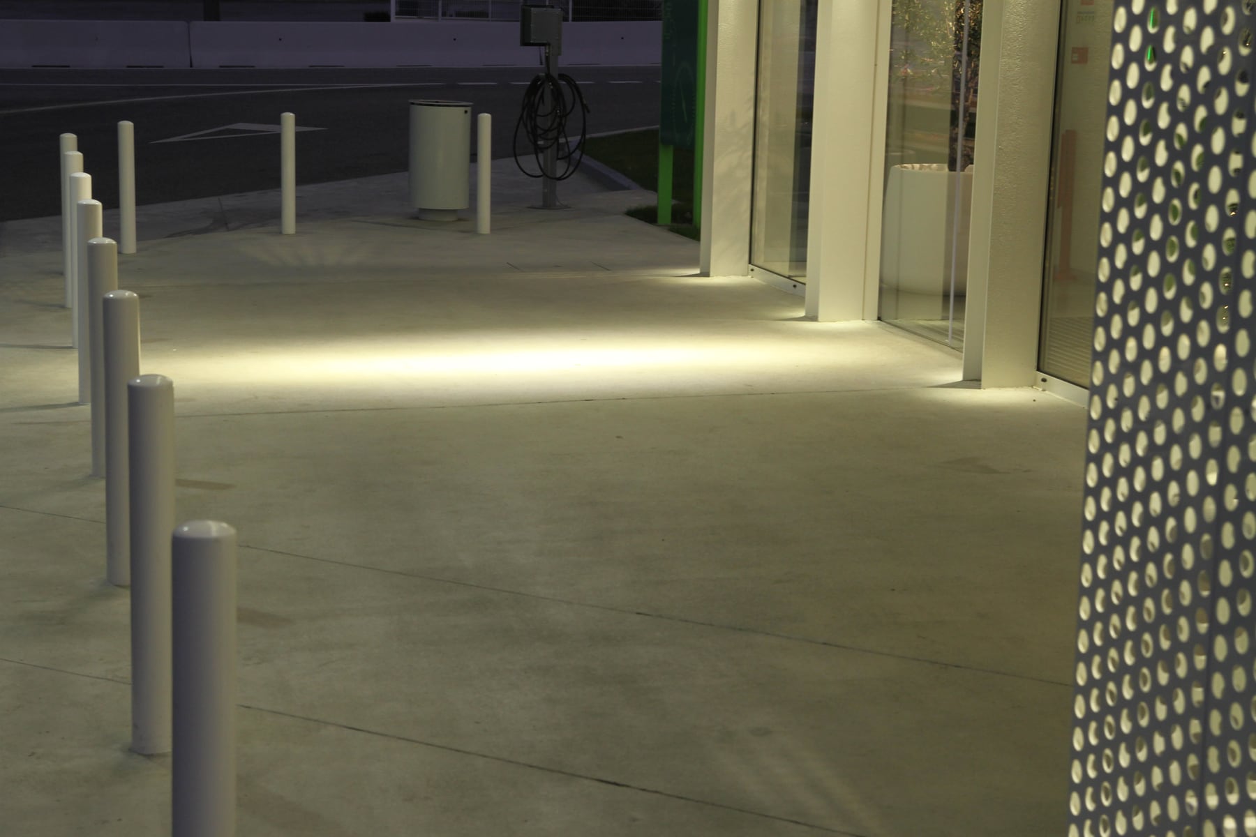The station forecourt at night, a line of light grazing the pavement beside the shop window and the white bollards lined up at left.