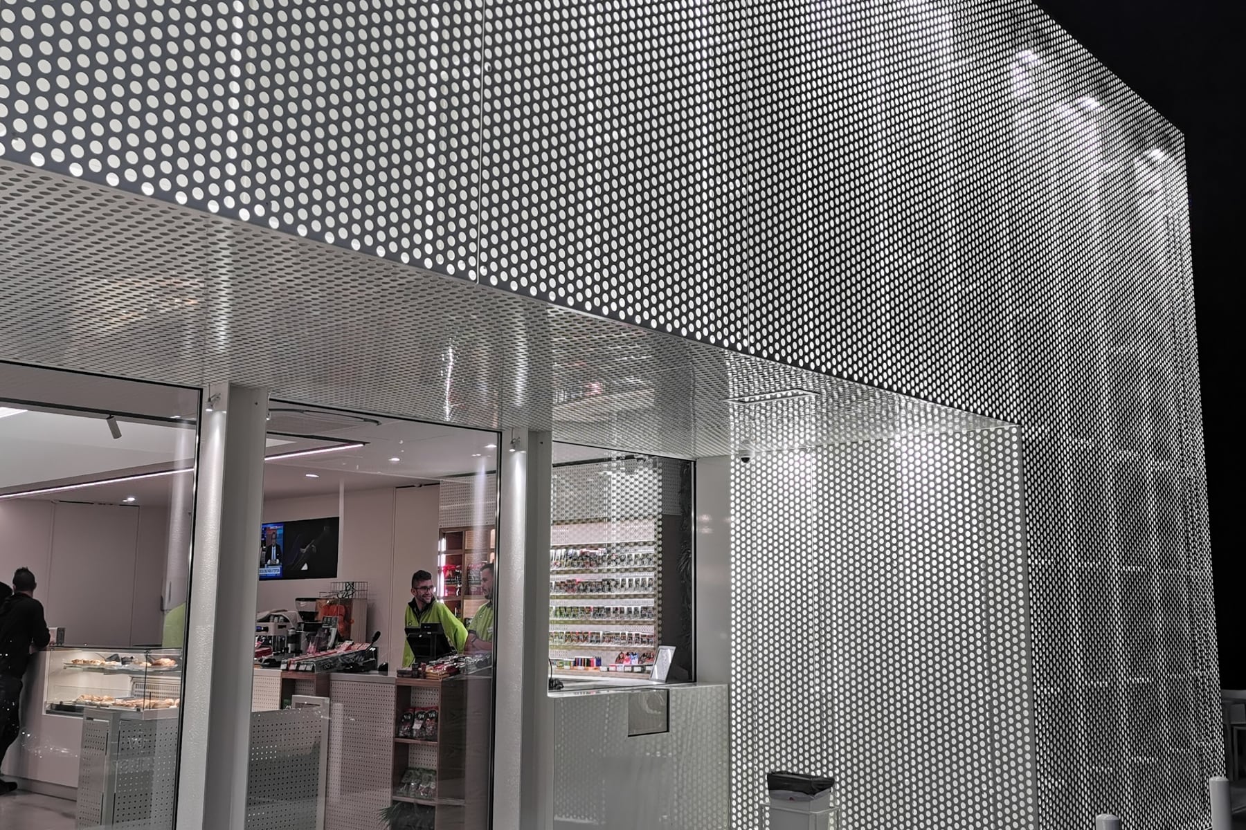The perforated metal skin seen close up, the shop lit behind it and the floodlights grazing the sheet from above.