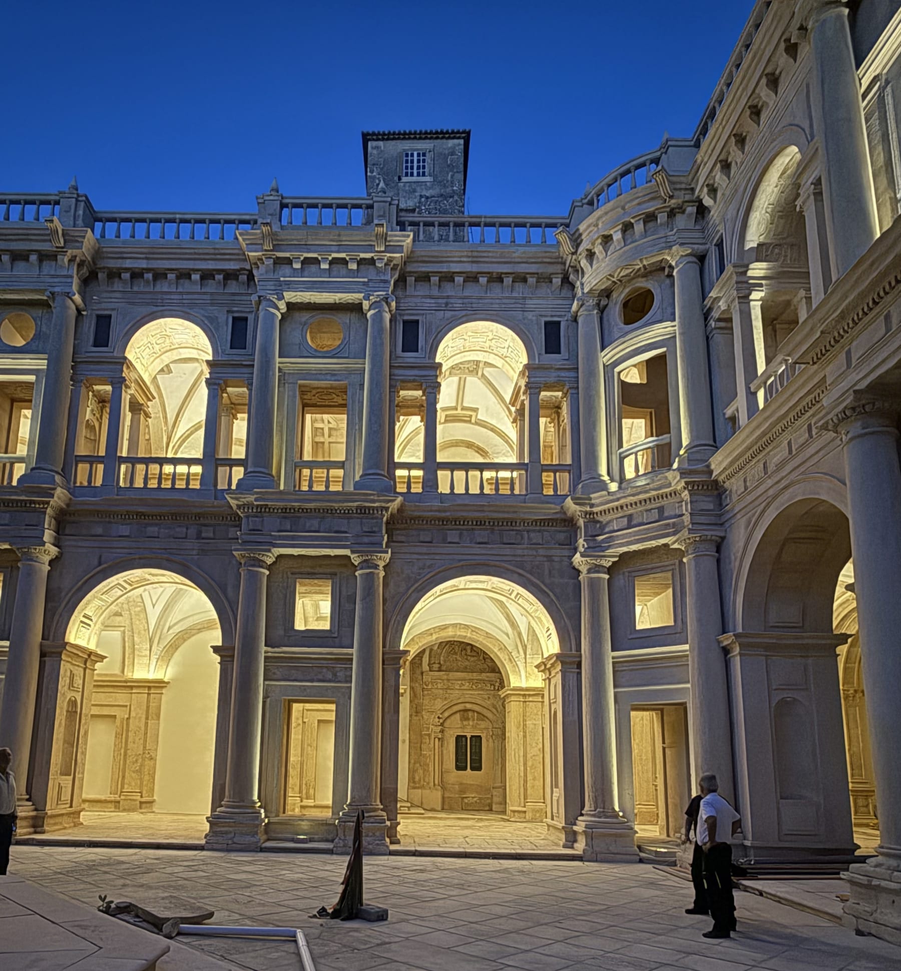The Claustro de Dom João III at the Convento de Cristo at dusk, both orders of arcading lit from within against the blue sky.