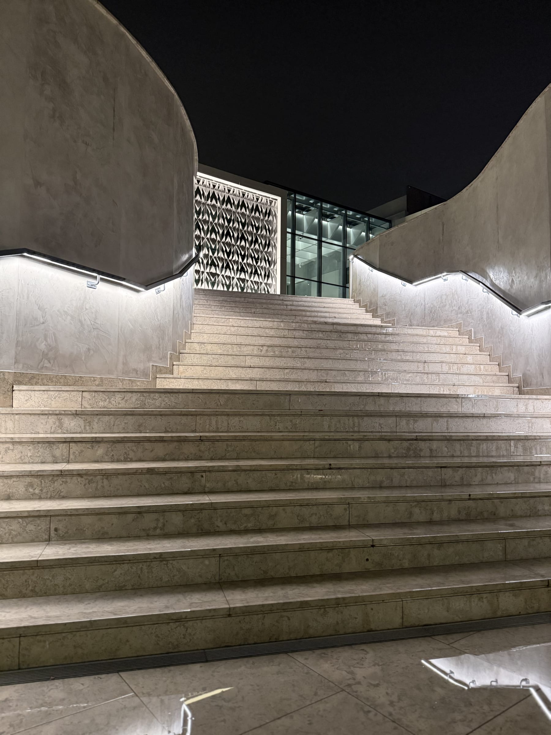 The exterior approach stair to the hotel and the museum at night, the handrails lit from within drawing the climb against the ceramic panel beyond.