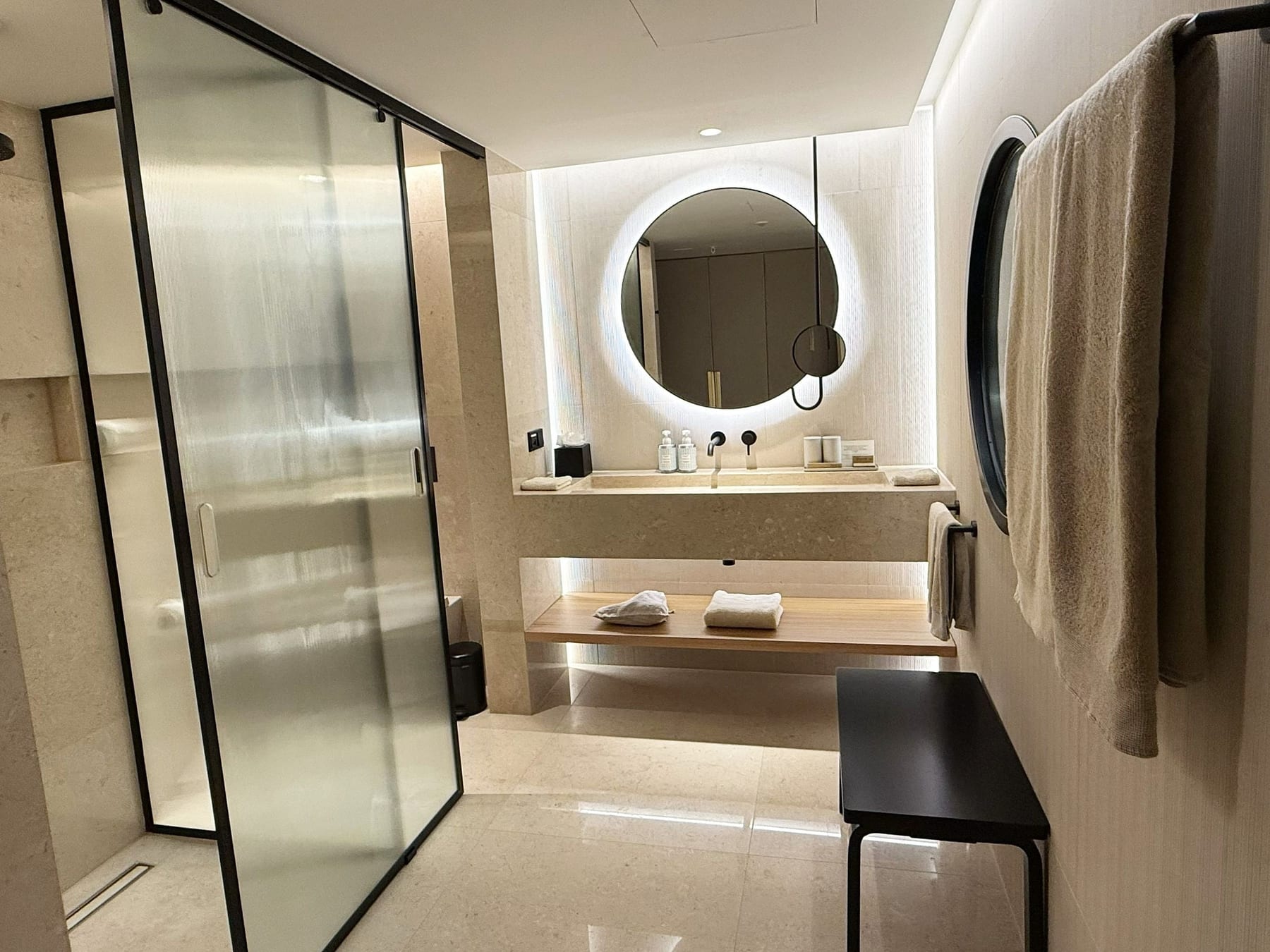 The bathroom of a new-wing room, the fluted glass shower at the left, the backlit round mirror over the stone basin and the timber shelf washed from below.