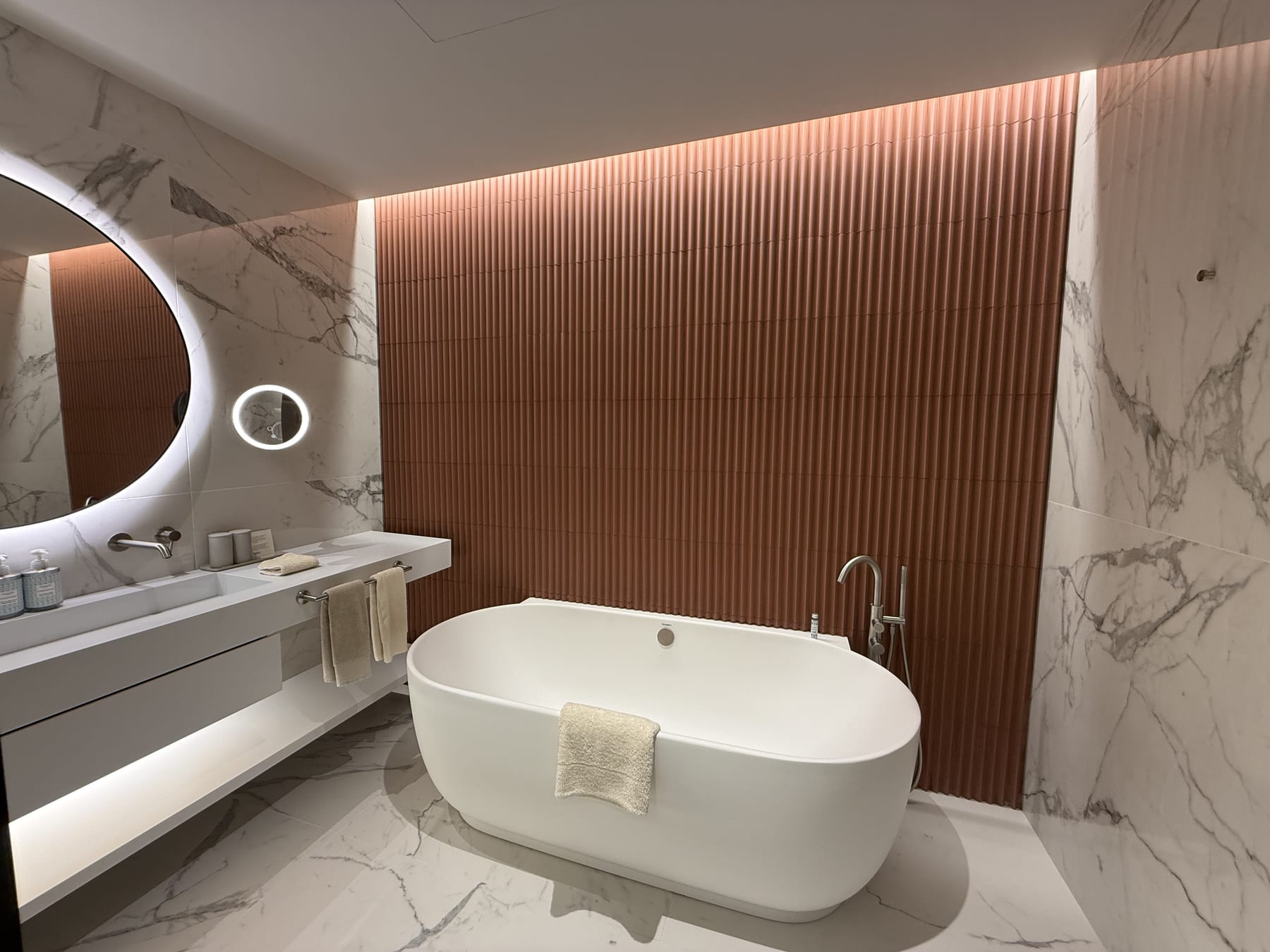 The bathroom of the same room, the fluted terracotta tile grazed with light and the round mirror backlit.