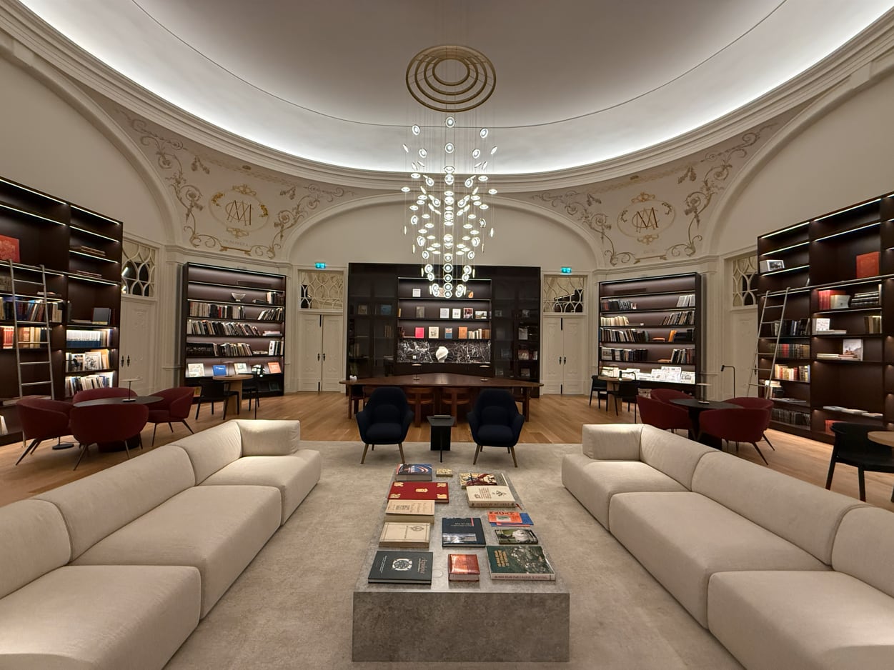 The hotel library under its cove-washed dome, linear light built into the shelving, the crystal chandelier at the centre and the palace monograms on the spandrels.