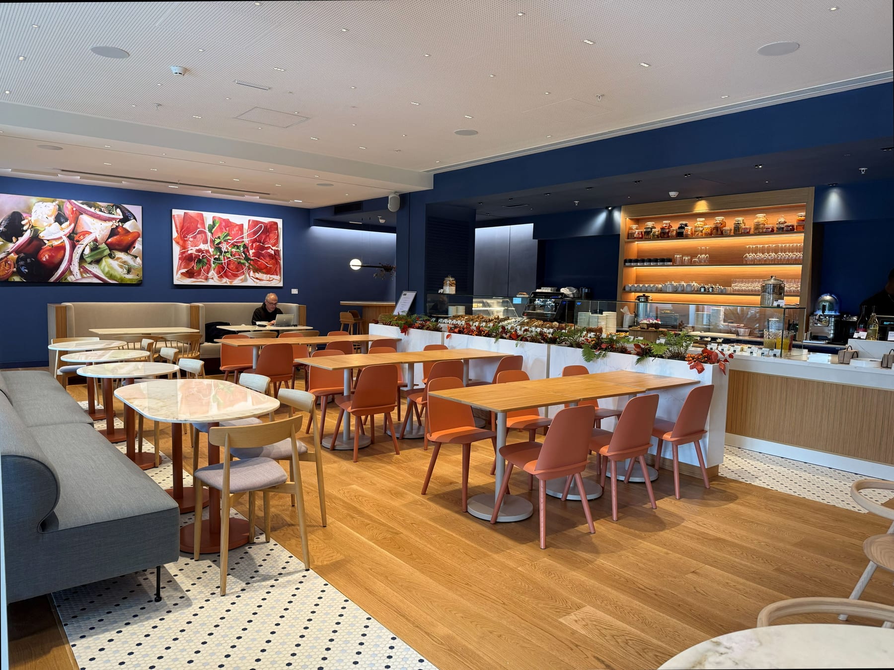 The museum cafeteria, the counter shelving lit from within, the blue wall beyond and terracotta chairs on the timber floor.
