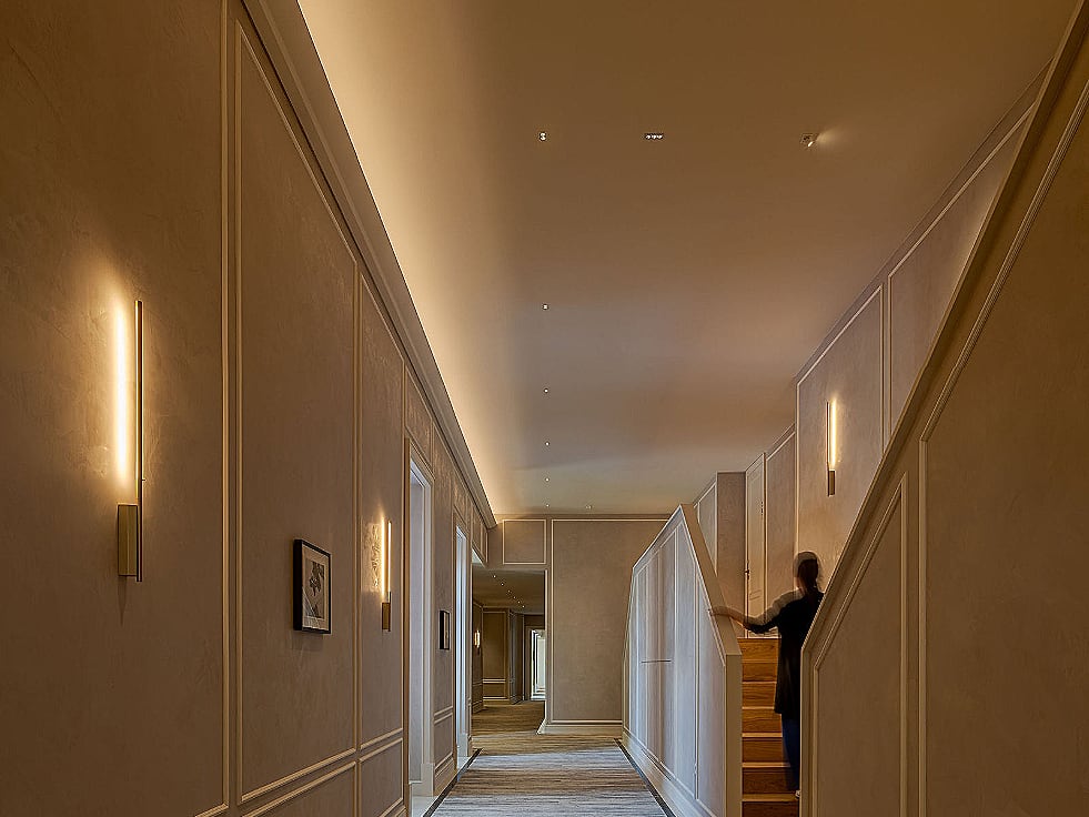 A guest floor corridor, vertical linear wall lights on the plastered wall and a cove washing the ceiling to the far end.