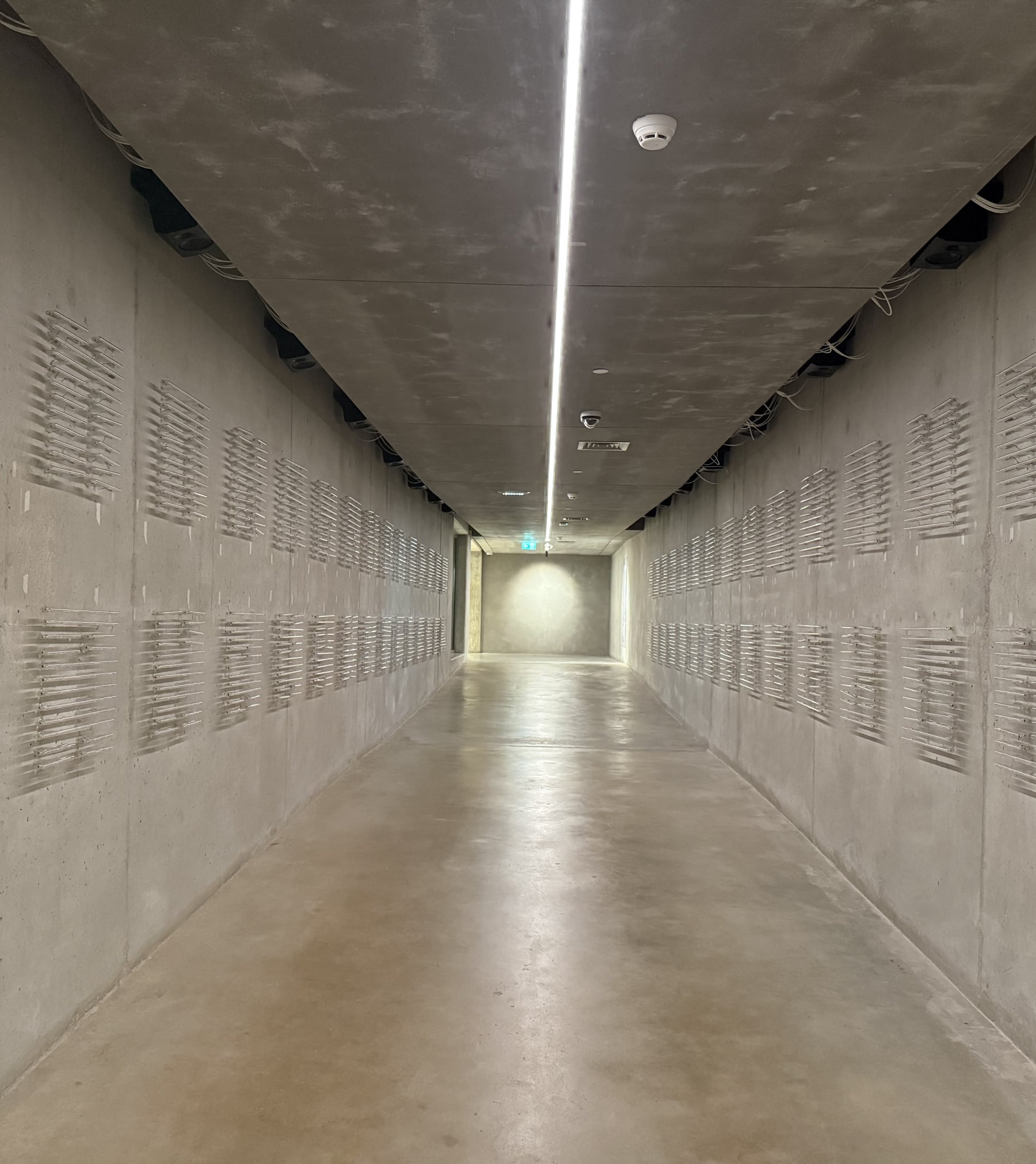The approach corridor from the car park, a continuous line of light on the ceiling pulling the perspective and the names cut into the concrete walls.