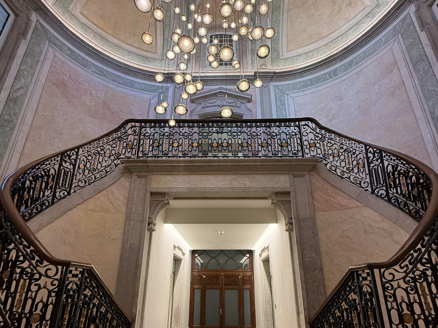 The monumental staircase seen from below, its two flights opening under the cascade of globes with the lit enfilade beyond.