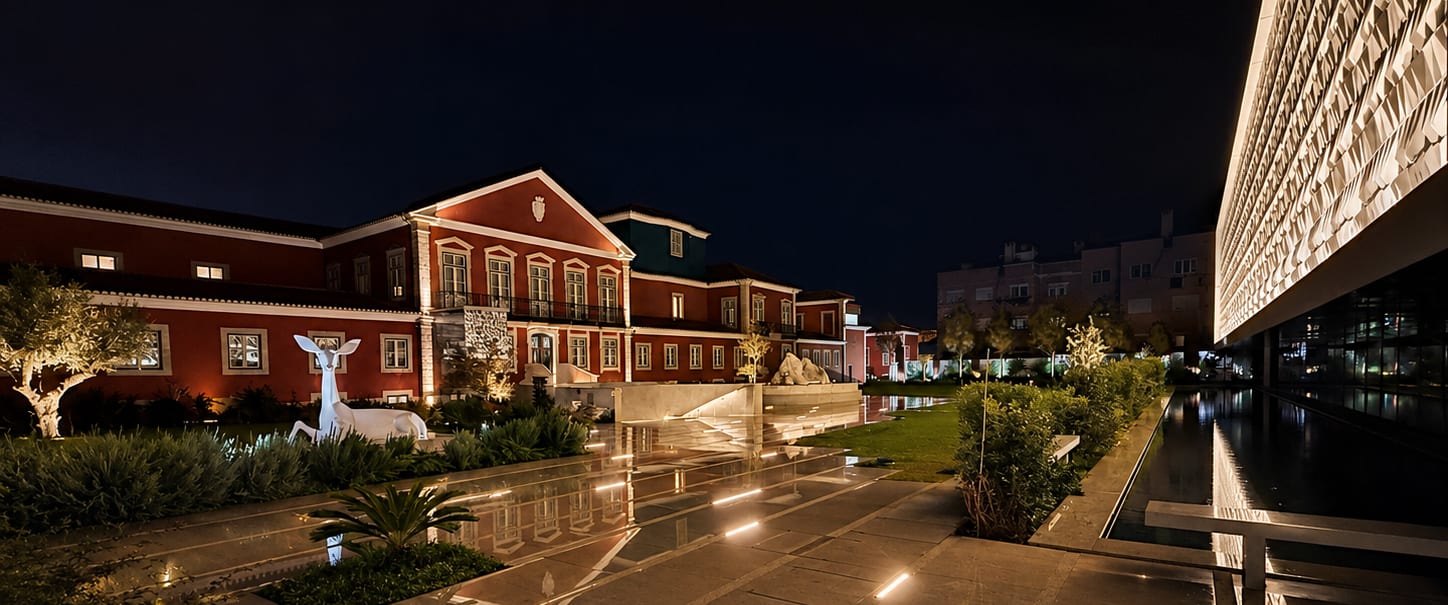 The MACAM complex at night: the palace facade grazed with light and returned by the water mirror, light lines set into the paving, and at the right the perforated ceramic facade of the new wing.