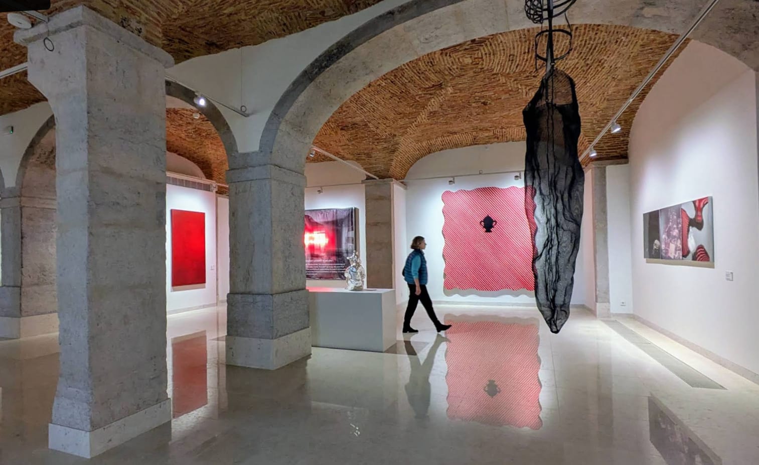 The gallery under the brick vaults, the stone piers taking a grazing light, each work held by its own spot and the tulle sculpture hung from the vault; the polished floor returns the whole room.