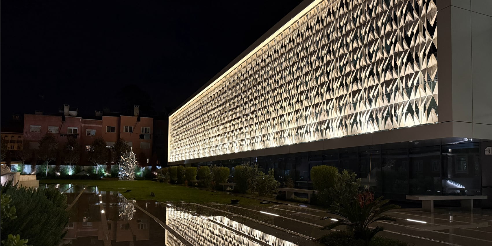 The galleries from outside at night: the perforated ceramic facade lit from within and returned by the water mirror.
