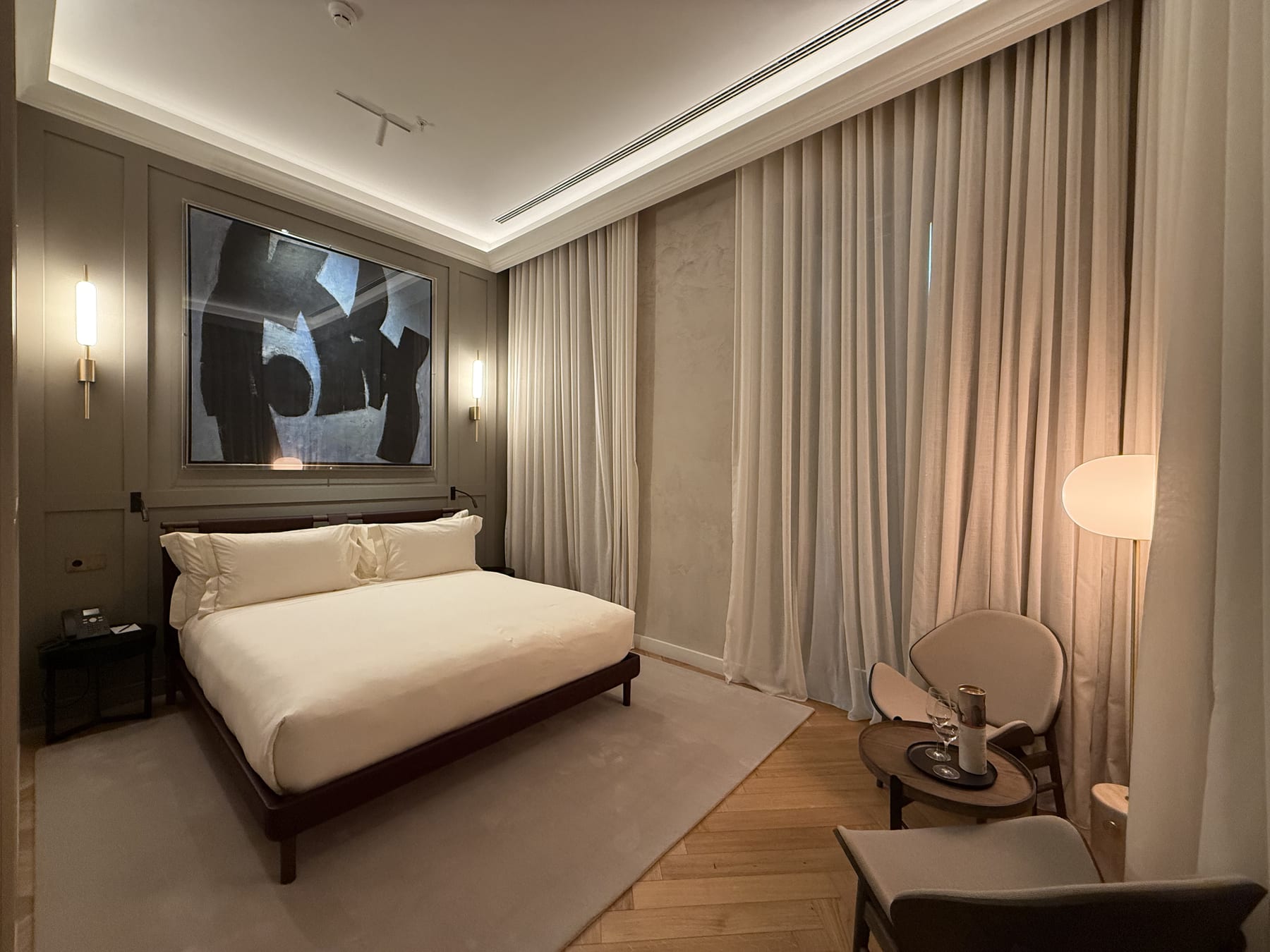 A room in taupe, vertical wall lights flanking the painting and the cove washing the ceiling above linen curtains.