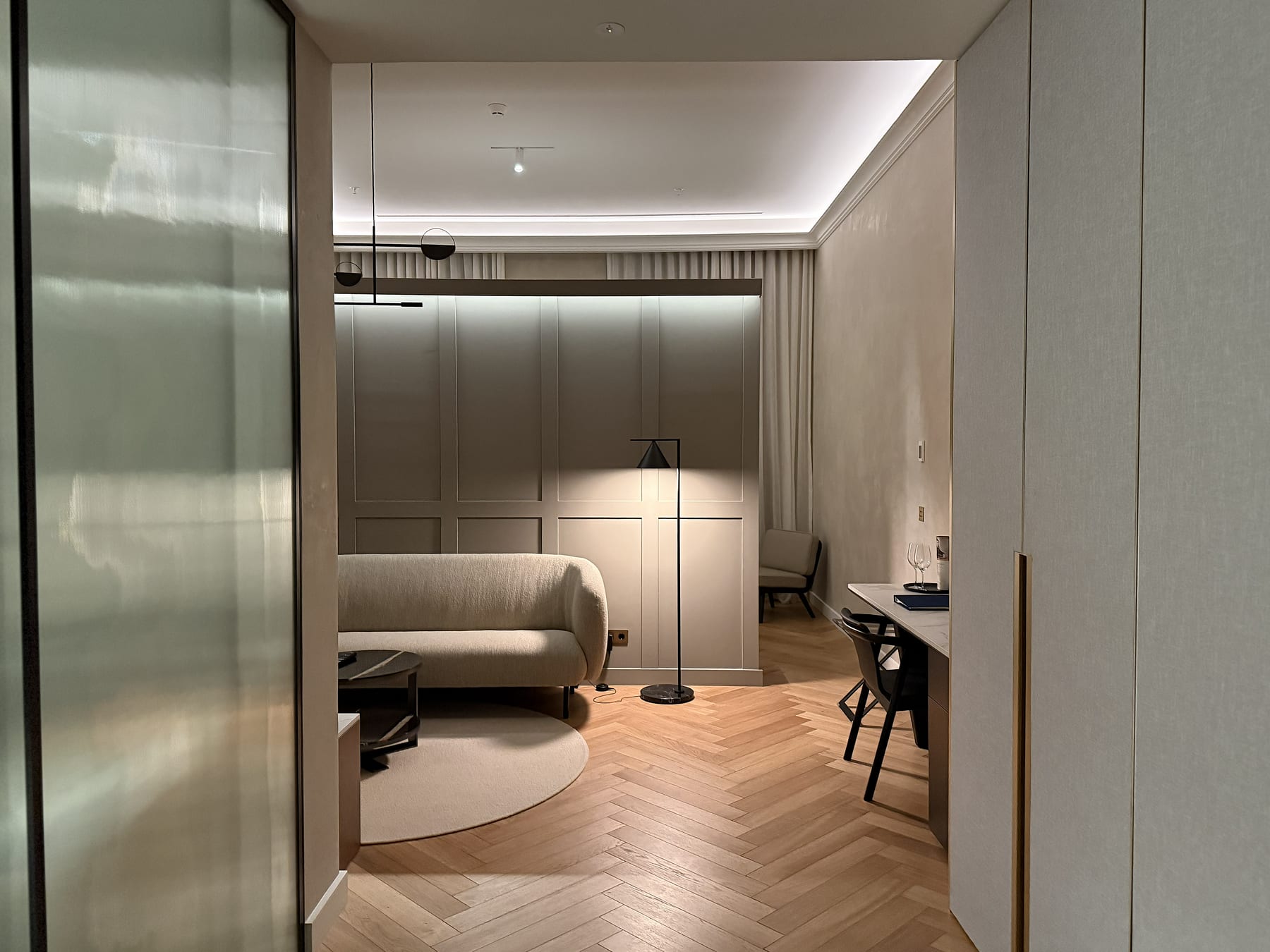 A guest room seen through the frosted glass partition, the ceiling washed by a cove and a floor lamp over herringbone parquet.