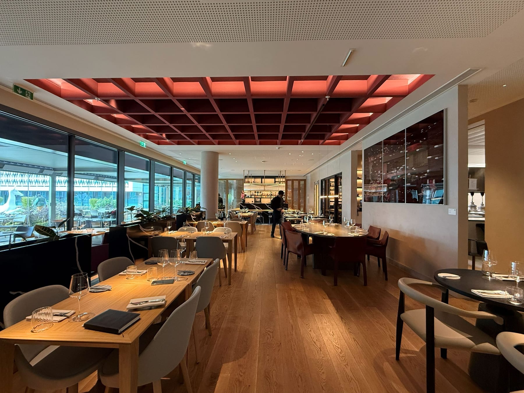 The restaurant dining room, the coffered ceiling lit pink over the laid tables, the counter at the far end and the glazed facade to the left.