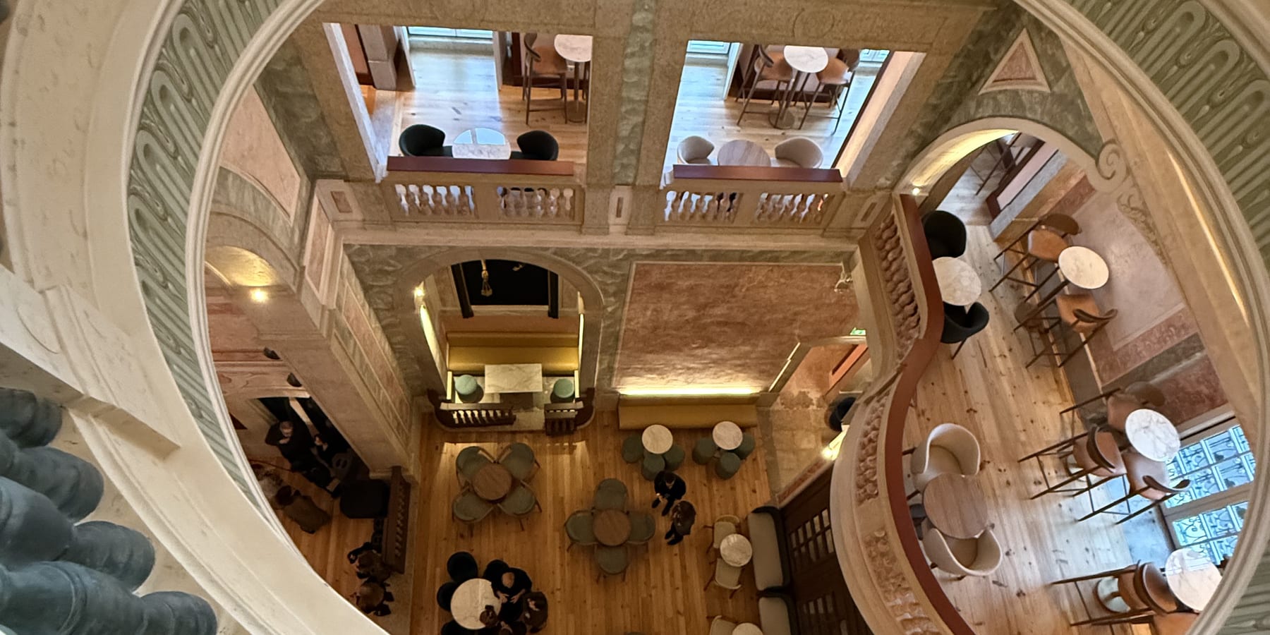 The MACAM atrium seen from above, from the piano nobile: the café tables on the timber floor, the bar at the right, and the stone arc of the opening sweeping across the frame.