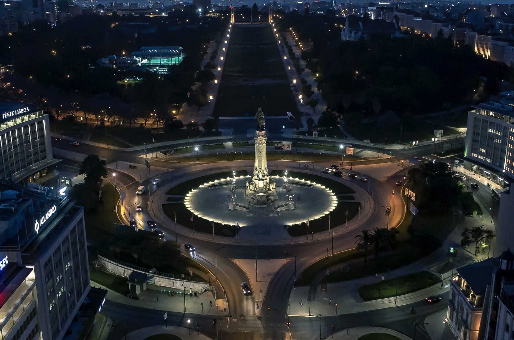 The Marquês de Pombal roundabout seen from above at dusk, the ring of light drawing the garden around the column and the Avenida climbing to the park.