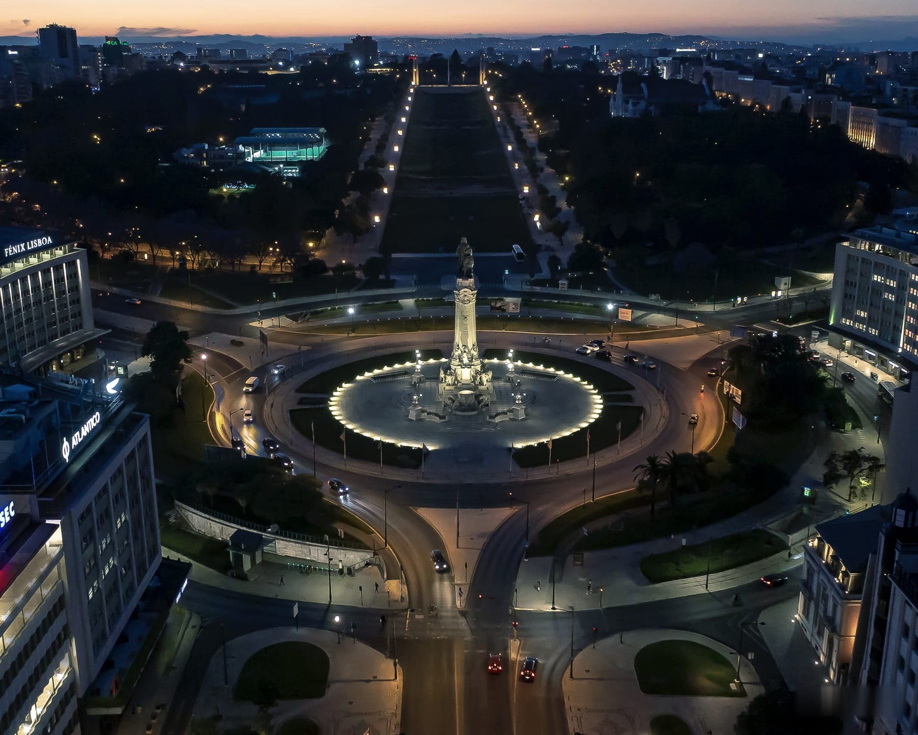 Aerial view of the Marquês de Pombal roundabout at dusk, the ring of light drawing the garden around the monument, with the Avenida da Liberdade and Parque Eduardo VII opening out behind.