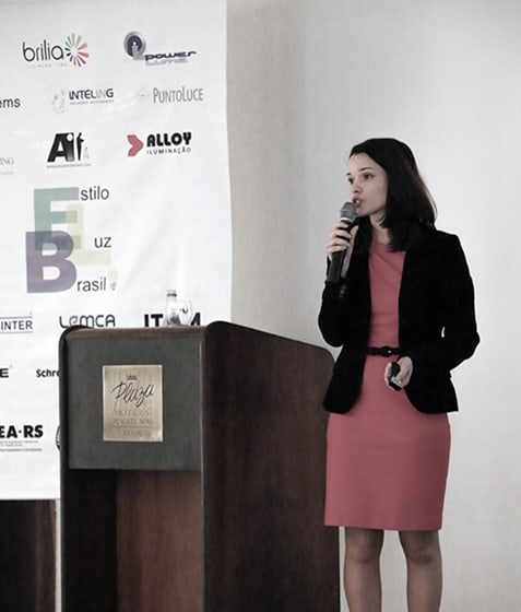 Diana Delnegro speaking at the lectern at Estilo Luz Brasil, in Porto Alegre, the sponsors’ board behind her.