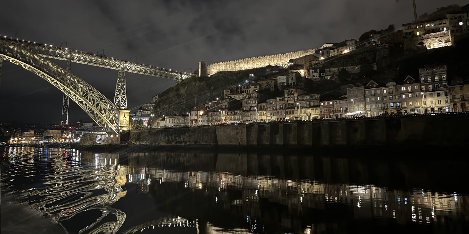 The Fernandine Wall lit above the Ribeira hillside, seen from the Douro at night, with the Luís I bridge to the left and the lights reflected in the water.