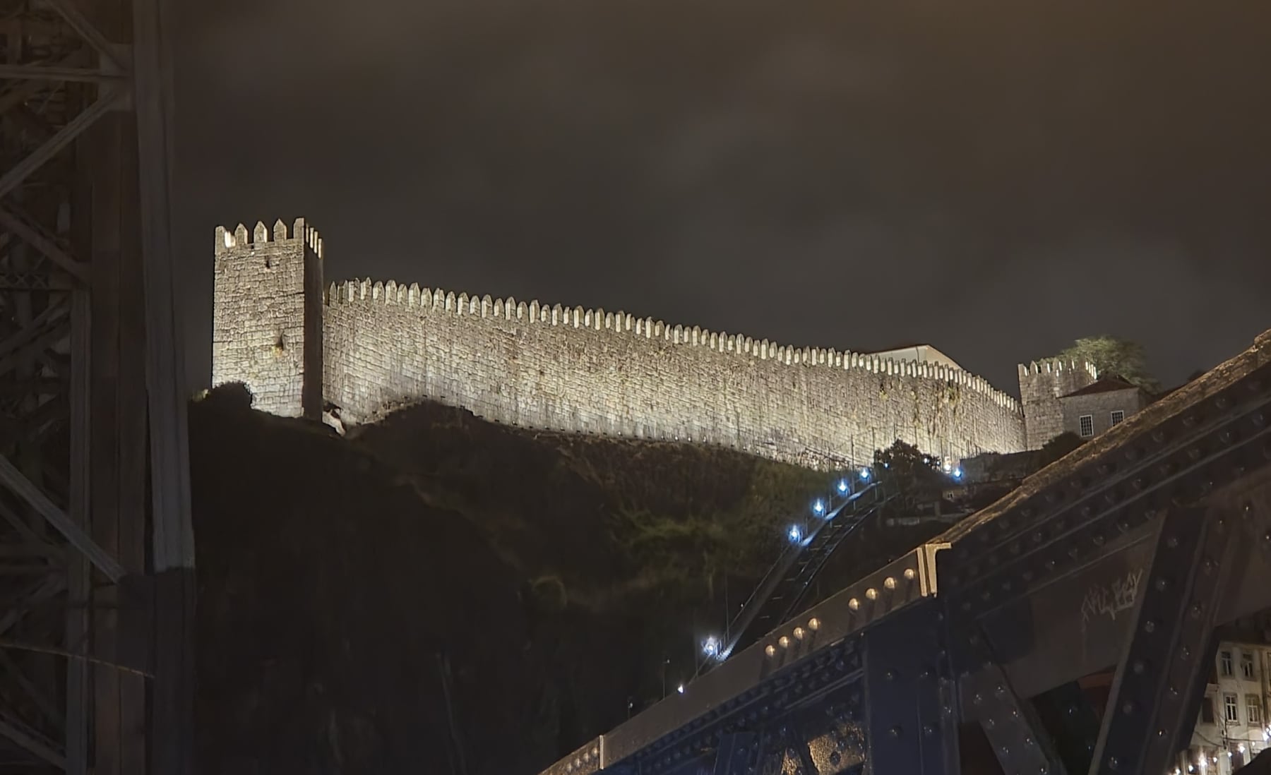The Fernandine Wall seen from under the Luís I bridge, the stone curtain lit as it climbs the hillside and the merlons cut against the sky.