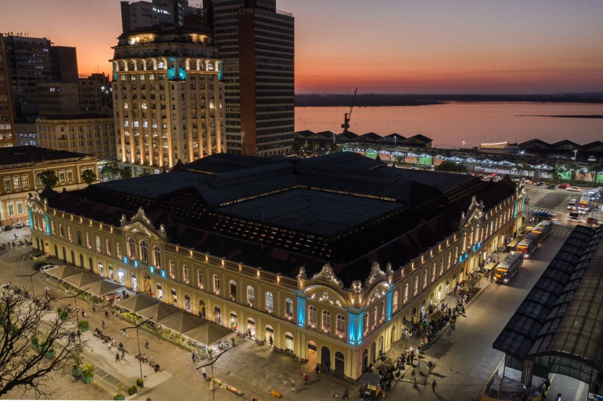 The Mercado Público of Porto Alegre lit at dusk, seen from above, the facades in warm light and the pilasters marked in blue, the Guaíba beyond.